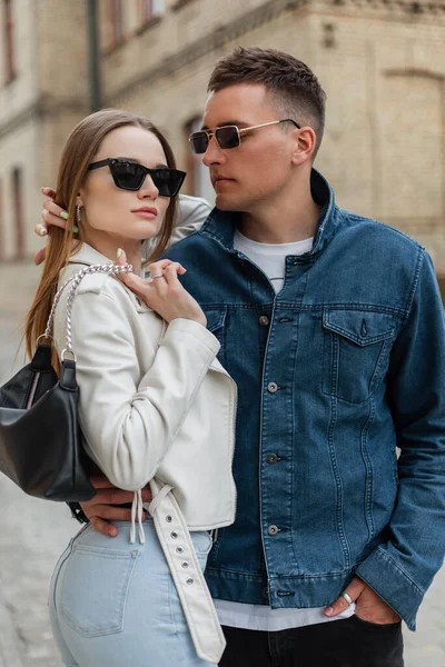 Trendy fashionable young beauty couple woman and man with stylish sunglasses in denim and leather jacket with bag poses and walks on the street