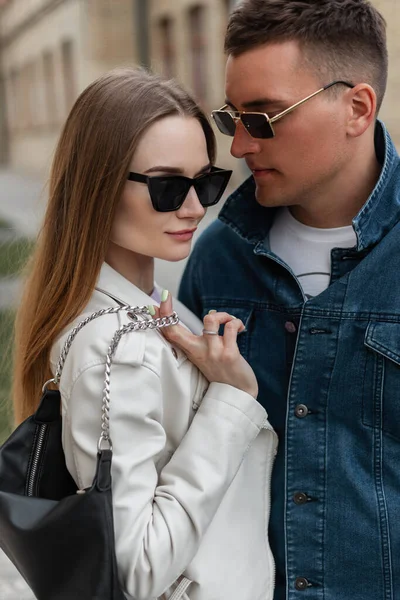 Stylish beautiful street couple of models with trendy glasses in trendy casual clothes with a jeans jacket and a leather jacket with a bag are standing in the city. Trendy woman and handsome man