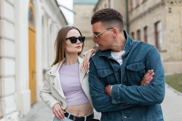 Beautiful young woman model and a hipster man in fashionable denim clothes with sunglasses are walking in the city. Urban fashionable couple outdoors