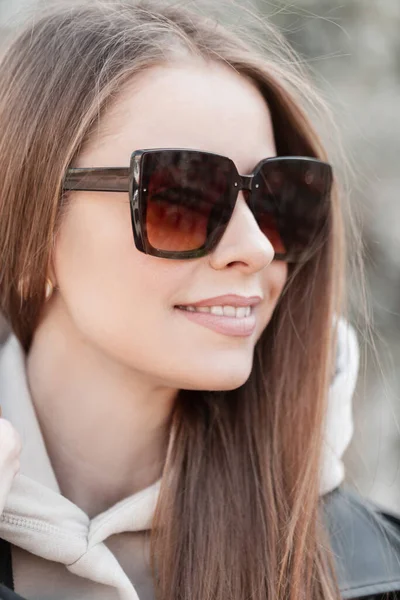 Women's happiness portrait of a beautiful stylish smiling girl model with fashionable vintage sunglasses in spring casual outfit with a leather jacket and hoodie walking on a sunny day in the city