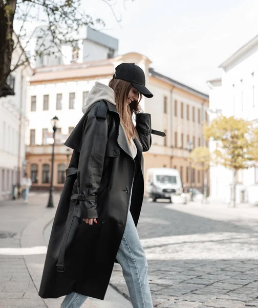 Stylish beautiful trendy hipster woman with a cap in trendy black clothes with a fashion long black leather coat, hoodie and jeans is walking in the city