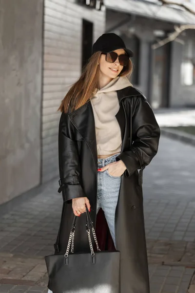 Happy urban fashion girl with a smile with sunglasses and a fashionable cap in a stylish leather coat with a hoodie, jeans and a bag walking on the street near the building