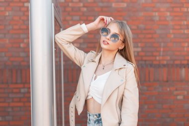 Women's street portrait of a beautiful stylish pretty girl in fashionable outfit with fashion sunglasses stands near a brick building in the city