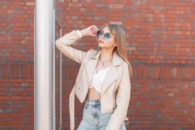 Beautiful zoomer fashionable young woman model in a white leather jacket with a top and jeans wears sunglasses near a brick wall in the city. Urban female youth street style clothes look