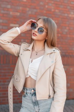 Stylish beautiful zoomer girl in fashionable clothes with a leather white jacket and top with vintage jeans standing near a brick wall and wearing round  sunglasses