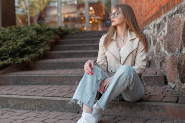 Stylish beautiful young woman with sunglasses in fashionable casual clothes with a leather jacket, top and jeans with white sneakers sits on the steps near a vintage brick building shopping mall.