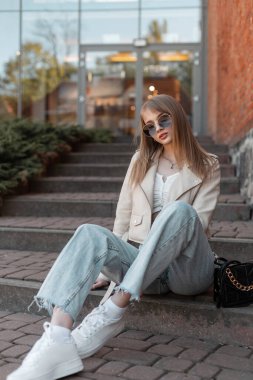 Casual trendy pretty woman with sunglasses in fashionable white leather jacket and vintage jeans with sneakers and bag sits on the steps near a brick building wall