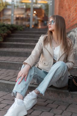 Fashionable beautiful young hipster girl with sunglasses in fashion casual outfit with leather jacket, jeans and white sneakers with a purse sits on the steps outside the mall in the city