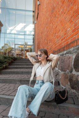 Trendy fashionable beauty girl model with fashion sunglasses in casual clothes with leather jacket, blue vintage jeans and purse sits and poses on the steps near a red brick mall
