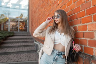 Women's fashion style of outfit. Beautiful young woman in a fashionable leather jacket, jeans with a bag wears trendy round sunglasses and stands near a red brick wall outside a shopping mall.