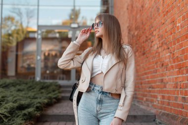 Pretty beautiful fashion young girl in a trendy leather white jacket with fashionable vintage high-waisted jeans wears stylish sunglasses and walks in the city