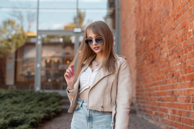 Stylish fashionable beautiful girl with glasses in a fashionable leather jacket with a top and high waist jeans walking on the street near the vintage red brick shopping mall. Female style fashion