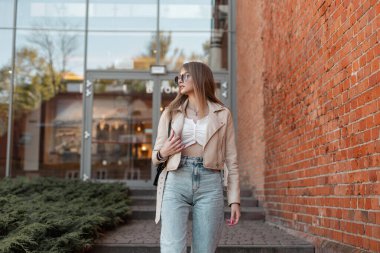 Beautiful young fashionable hipster zoomer woman in a trendy leather jacket with a fashion high waist jeans and stylish cool sunglasses walking on the street near the shopping mall