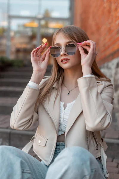 Cool pretty young zoomer woman with leather fashion white jacket and jeans wears a stylish sunglasses sits on the steps and rest near a vintage brick mall building