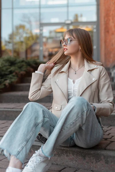 Fashion young woman model with sunglasses in leather white stylish leather and fashionable blue vintage jeans with sneakers sits on the steps near a mall