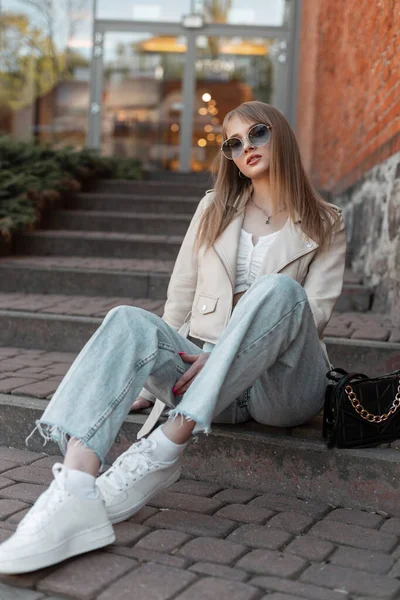 Beautiful fashionable hipster zoomer woman in fashionable street clothes with a white leather jacket, jeans and stylish sneakers with a purse is sitting on the steps in city near the shopping mall