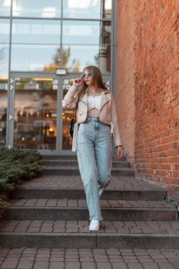 Fashionable young pretty woman with sunglasses in casual street outfit with leather jacket, jeans and white shoes walks near vintage brick building mall