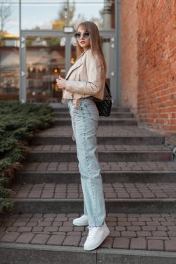 Stylish beautiful young woman with trendy sunglasses in a fashionable beige leather jacket with fashion vintage high waist jeans and white sneakers standing near the brick shopping mall