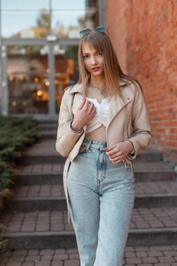 Cool hipster beautiful young stylish woman in fashionable clothes with a leather jacket and a top with vintage jeans walking near a shopping mall in the city