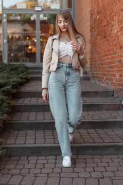 Stylish beautiful young woman in fashion clothes with fashion leather jacket, high-waisted jeans and white sneakers walks on the steps outside a vintage shopping mall