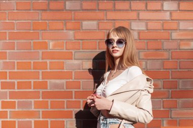 Stylish beautiful young girl model with fashionable sunglasses and fashionable clothes stands near a vintage brick wall in the city. Urban female style, fashion and beauty
