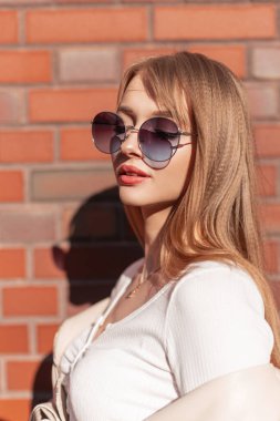 Stylish female city portrait of a beautiful young girl with fashionable round sunglasses in fashionable casual clothes walking on the street near a red brick wall on a sunny day, Fashion and beauty