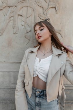 Fashionable beautiful young girl with a fresh face in fashionable casual urban clothes with leather jacket, jeans and top poses near a vintage column