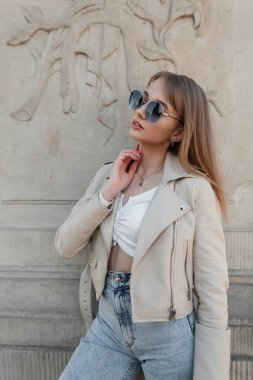 Fashion rock stylish beauty woman hipster with trendy sunglasses in fashionable leather jacket, jeans and white top poses near a vintage wall outdoors