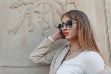 Sexy beautiful young woman hipster model with cool sunglasses in a fashion trendy white top with a leather jacket poses near a vintage wall outdoors