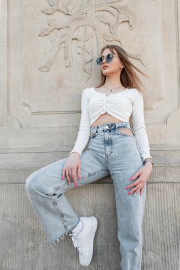 Cool stylish young pretty woman model with sunglasses in fashionable casual street clothes with a top, high waist jeans and white sneakers standing near a vintage column in a park in Poland