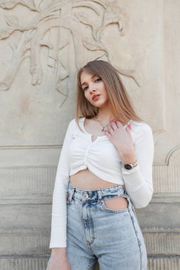 Pretty fashion young woman with stylish summer outfit with long sleeve top and vintage jeans stands near a vintage wall on the street