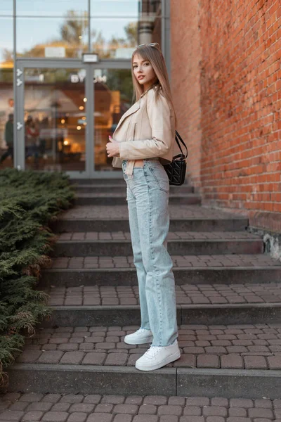 Stylish street pretty young woman in fashionable casual clothes with leather jacket, vintage high-waisted jeans, white shoes with black bag stands near a red brick shopping mall