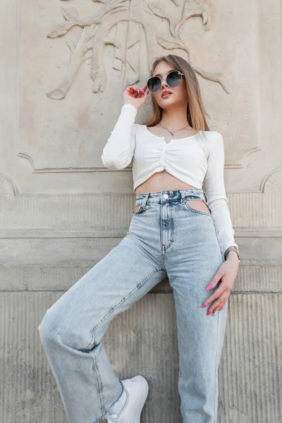 Stylish beautiful young girl model wears trendy sunglasses in a fashion white long-sleeved top with blue high-waisted jeans posing near a vintage wall. Urban female fashion and beauty