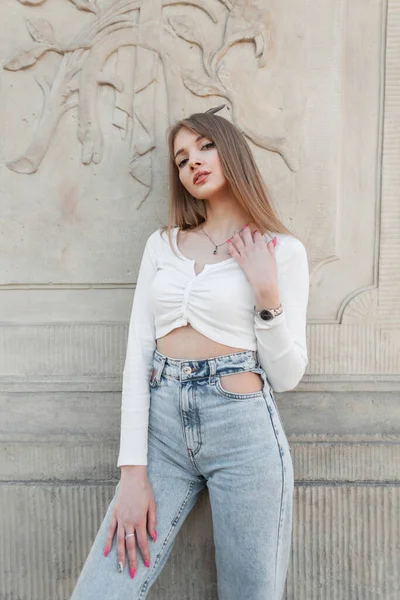 Beautiful girl with pretty Caucasian face in fashion white long sleeve top and high waist jeans poses near a vintage column on the street. Urban female streetwear look outfit