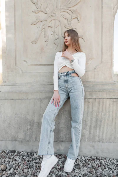 Fashion pretty woman hipster mode in trendy streetwear with stylish white long sleeve top, vintage height waisted jeans with white sneakers stands near a vintage column outdoors