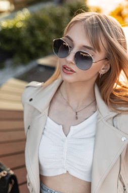 Beautiful female portrait of a beautiful young woman with cool round glasses wearing fashionable clothes with a leather rock jacket and top sitting in the city at sunset. Urban style outfit