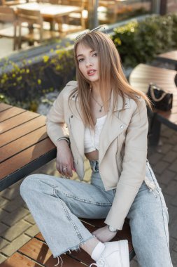Glamorous pretty woman hipster model with fashion casual rock style with leather jacket and vintage blue jeans sits on bench on the street