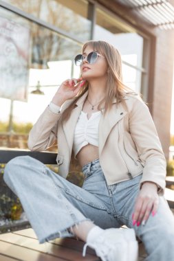 Fashionable beautiful young street hipster woman in fashion rock clothes with a stylish leather jacket, top, and vintage blue jeans with white sneakers sitting and relaxing in the city at sunset