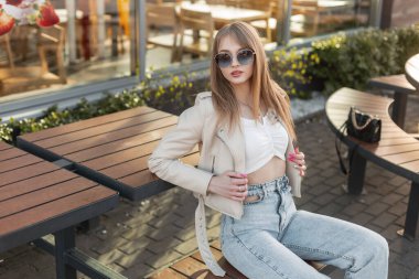 Fashionable beautiful stylish hipster girl model with round  sunglasses in a fashion leather rock jacket with a top and vintage blue jeans and a bag sitting on a wooden bench near a cafe at sunset