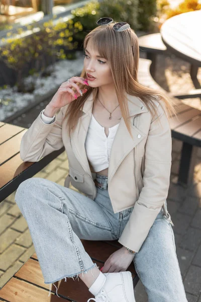 Fashionable beautiful fresh woman in fashion casual rock clothes with leather jacket, top and vintage blue jeans with white sneakers sits on a bench in the city