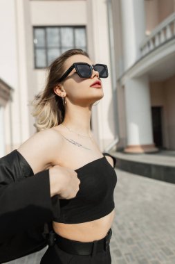 Stylish beautiful young girl with fashion black sunglasses in fashionable clothes with top, blazer and pants walking in the city near a vintage building
