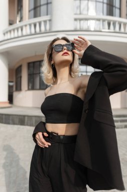 Fashion beauty glamour business woman in black elegant clothes with top, blazer and pants puts on a stylish sunglasses, walks and poses in the city near a vintage building