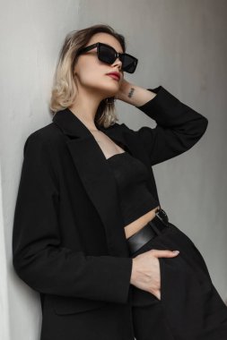 Trendy fashionable young woman model with short hair and sunglasses in fashionable business elegant black clothes stands near a white wall on the street