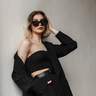 Fashionable young beautiful woman with cool modern sunglasses and short hair in business black elegant clothes with top, blazer and pants stands near white wall on the street.
