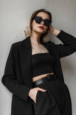 Fashionable business young beautiful woman model with trendy sunglasses in black fashion elegant blazer, top and pants stands near a white wall on the street