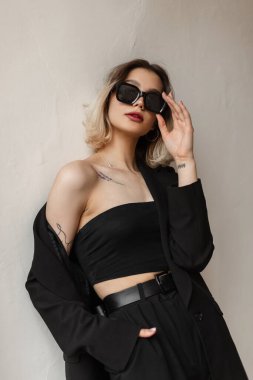 Cool fashionable vogue business woman in black stylish clothes with top, blazer and pants wears a fashion sunglasses and stands near a white wall