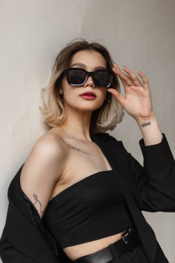 Fashionable beautiful young girl model with short hair in a fashionable black business elegant clothes with a top and a blazer is fixing modern sunglasses and standing near the white wall