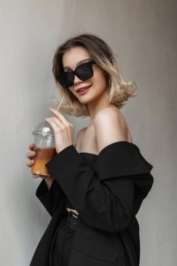 Cool fashionable beautiful young happy woman model with modern sunglasses in stylish black clothes with elegant fashion blazer drinks coffee and walks in the city