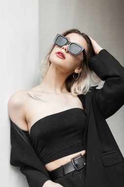 Fashionable beautiful business girl model with a short haircut and cool fashion sunglasses in fashionable elegant black clothes with a top and blazer standing near the white wall on the street