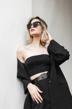 Elegant beautiful young woman with trendy cool sunglasses in fashion business black clothes with top, blazer and pants stands near a white wall on the street